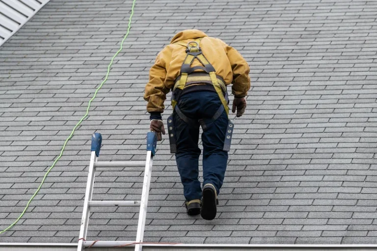 Worker in yellow hard hat and safety gear climbing ladder on residential roof